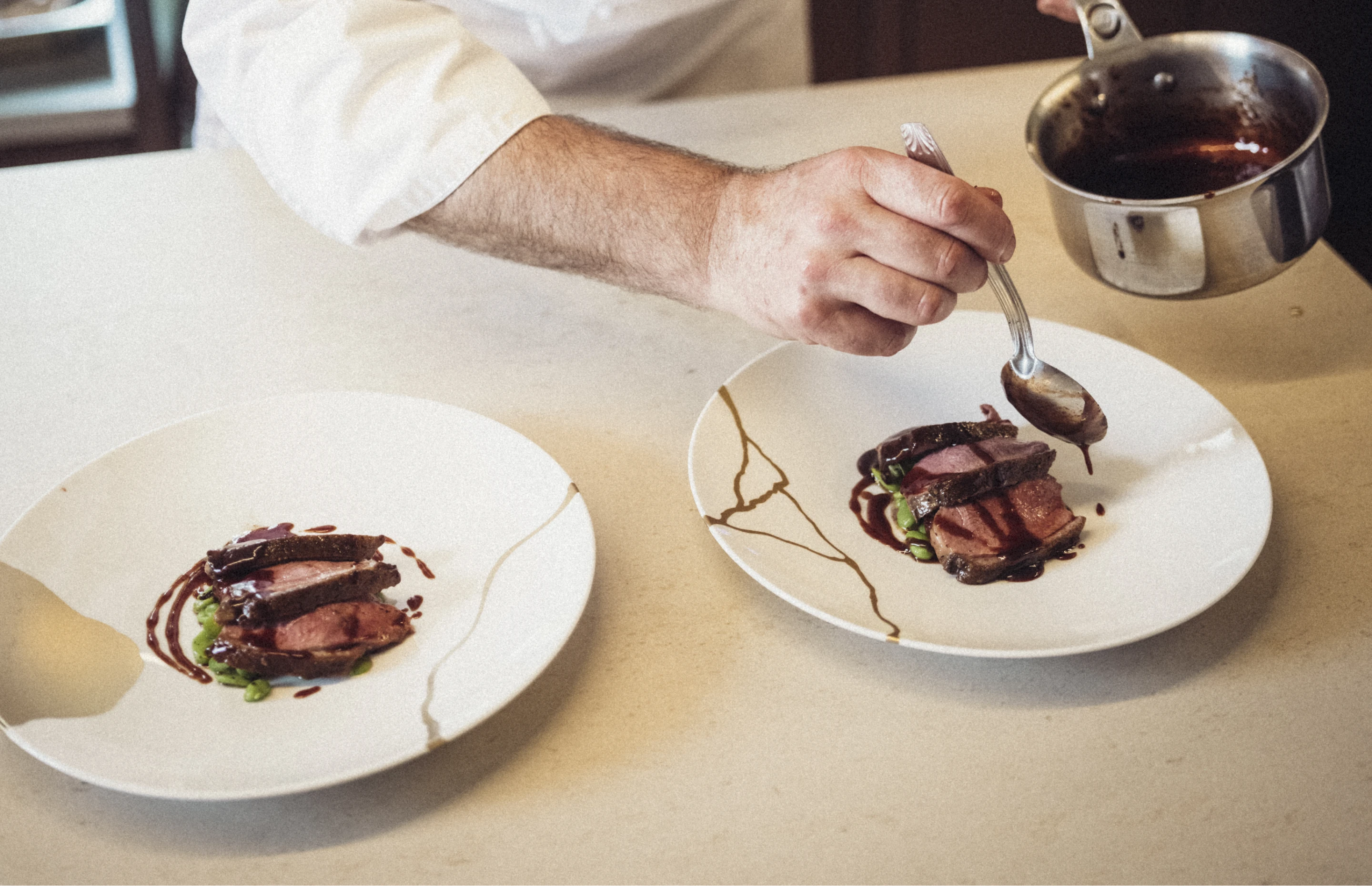 Un chef verse une sauce brune sur des tranches de viande sur une assiette blanche à côté d'une autre assiette similaire avec de la viande et des légumes verts.