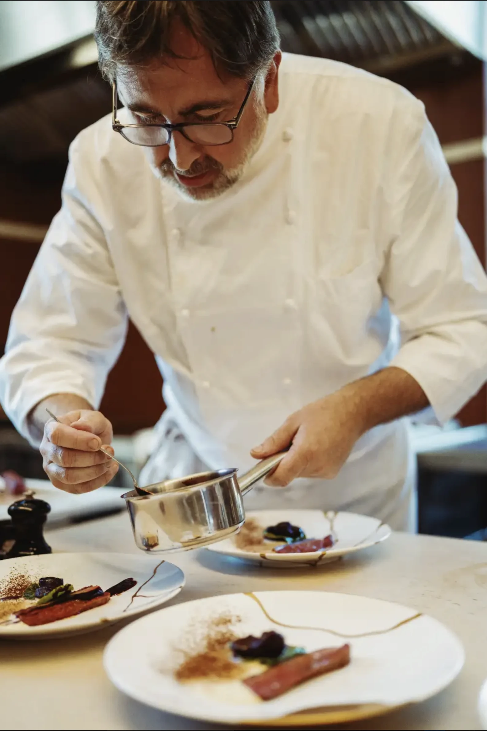 Chef concentré versant une sauce d'une petite casserole sur des plats élégants avec une présentation soignée.