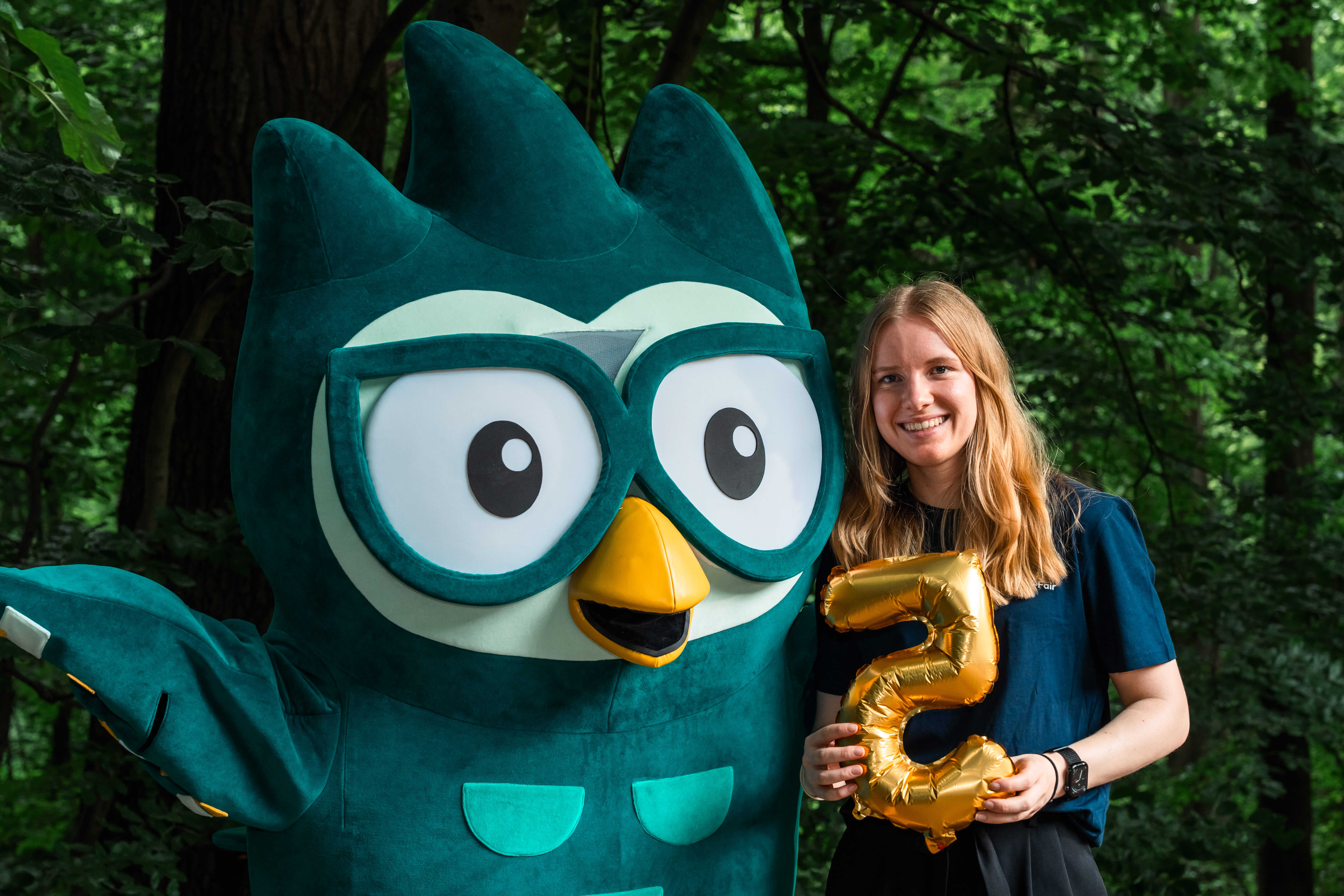 Junge Frau neben Eulen Maskottchen und goldner Luftballon Fünf in der Hand
