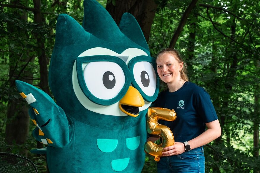 Junge Frau in Lern-Fair T-Shirt vor Wald mit Eulen Maskottchen