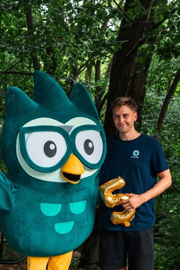 Junger Mann in Lern-Fair T-Shirt vor Wald mit Eulen Maskottchen