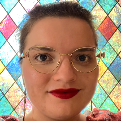 Eine junge Frau lächelt in die Kamera, mit Brille und vor buntem Glasfenster.