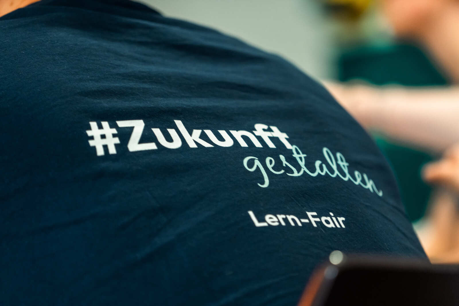 Der Rücken einer ehrenamtlichen Helferin fotografiert. Sie trägt das Lern-Fair T-Shirt mit dem Schriftzug #Zukunt gestalten.