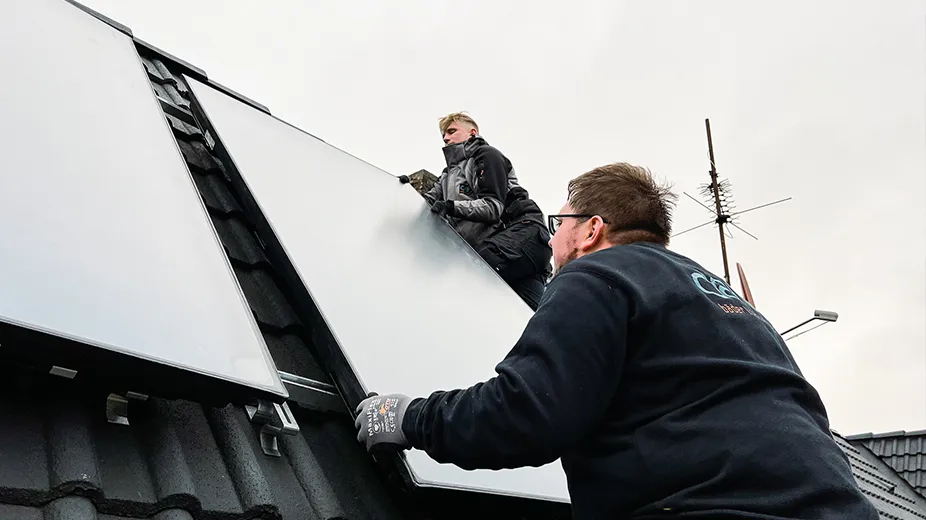 Zwei Männer installieren Solarpaneele auf einem schrägen Dach bei bewölktem Himmel.