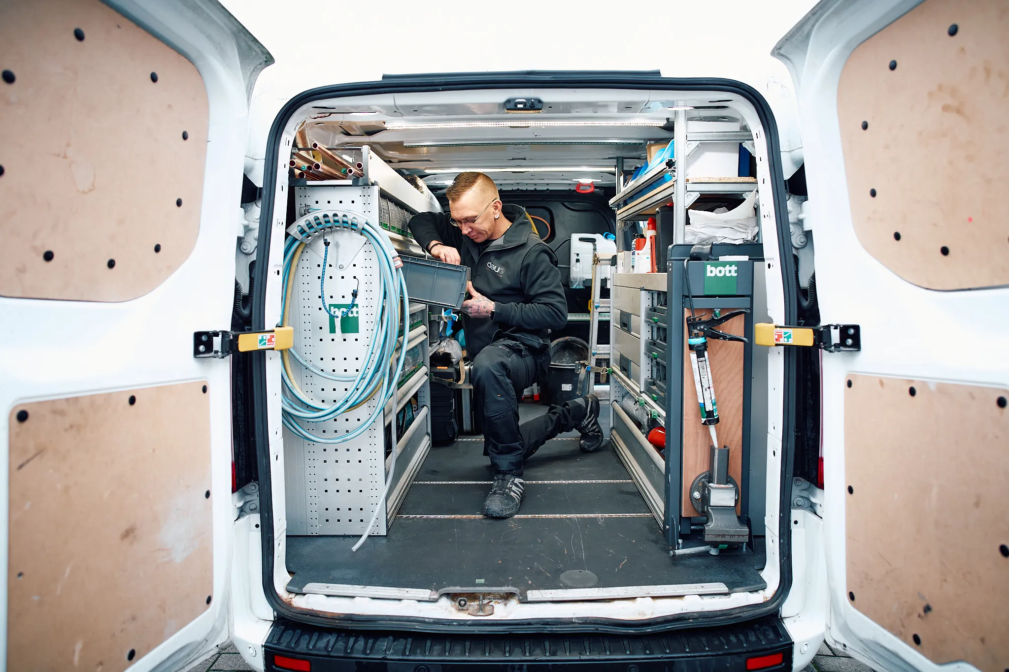 Handwerker kniet im geparkten weißen Transporter neben Werkzeugsystemen und organisiert Material.