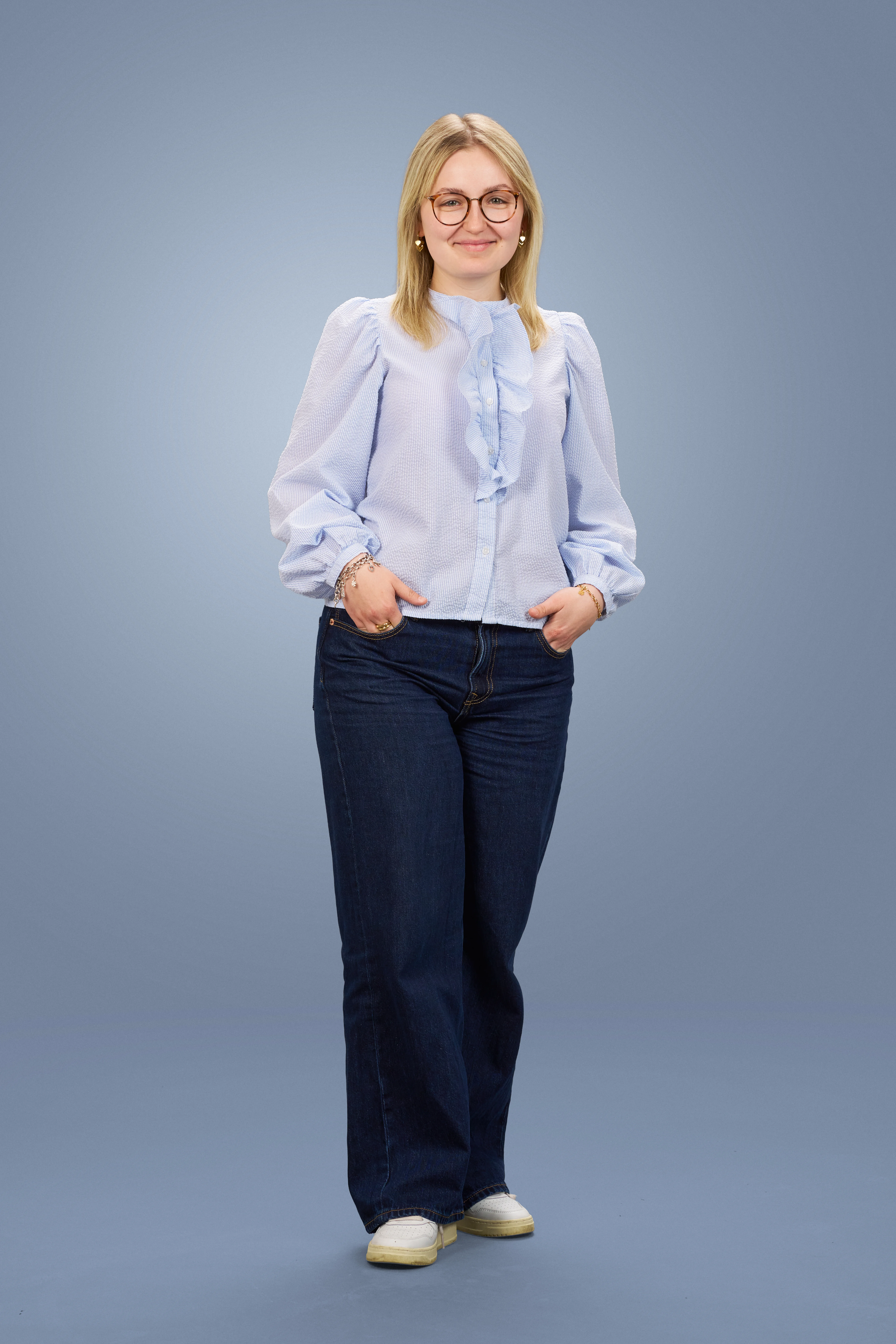 Junge Frau mit blonder Haarfarbe, Brille, hellblauer Bluse mit Rüschen und blauen Jeans lächelt.