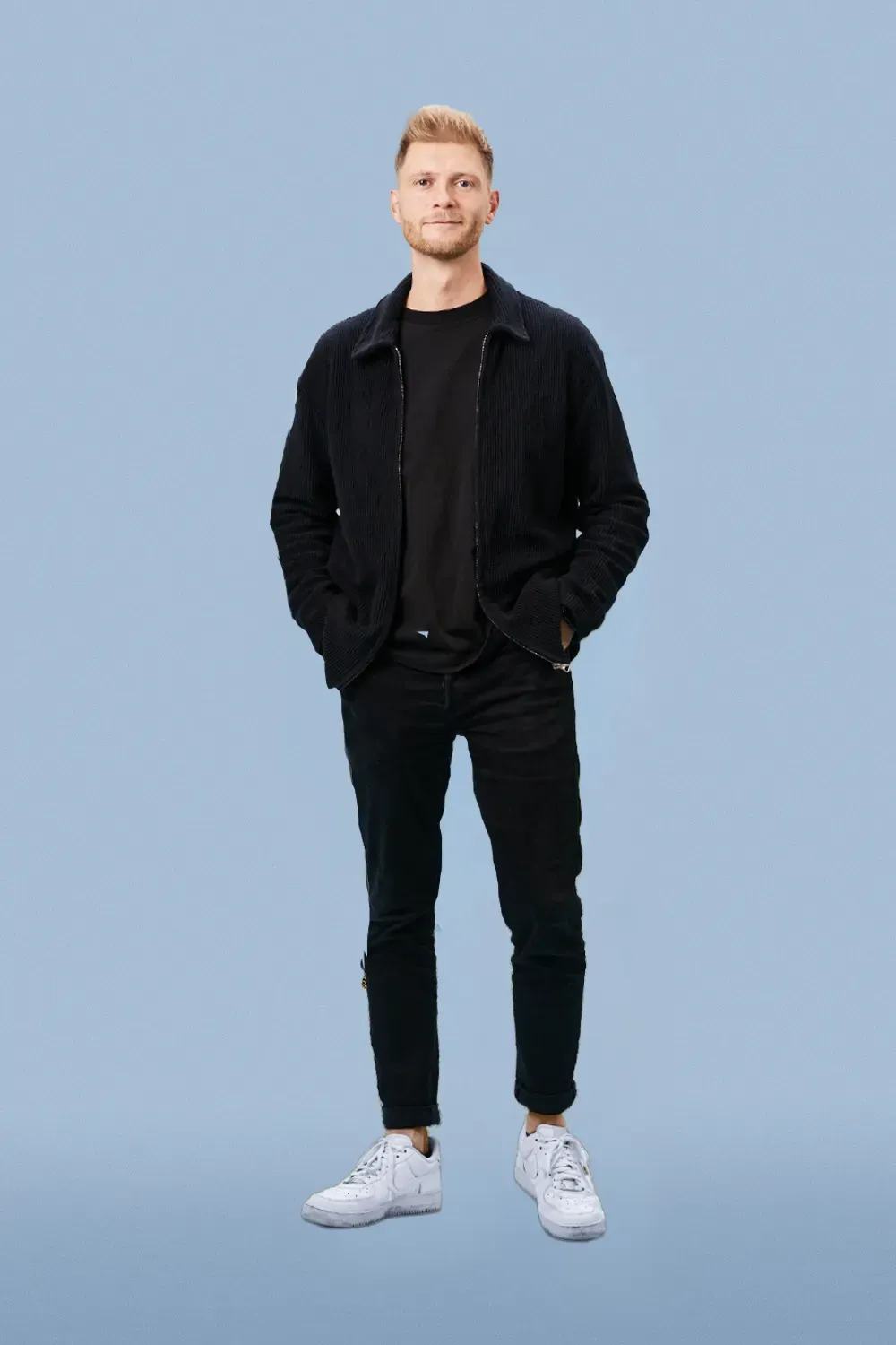 Junger Mann steht vor blauem Hintergrund, trägt schwarze Kleidung und weiße Turnschuhe.