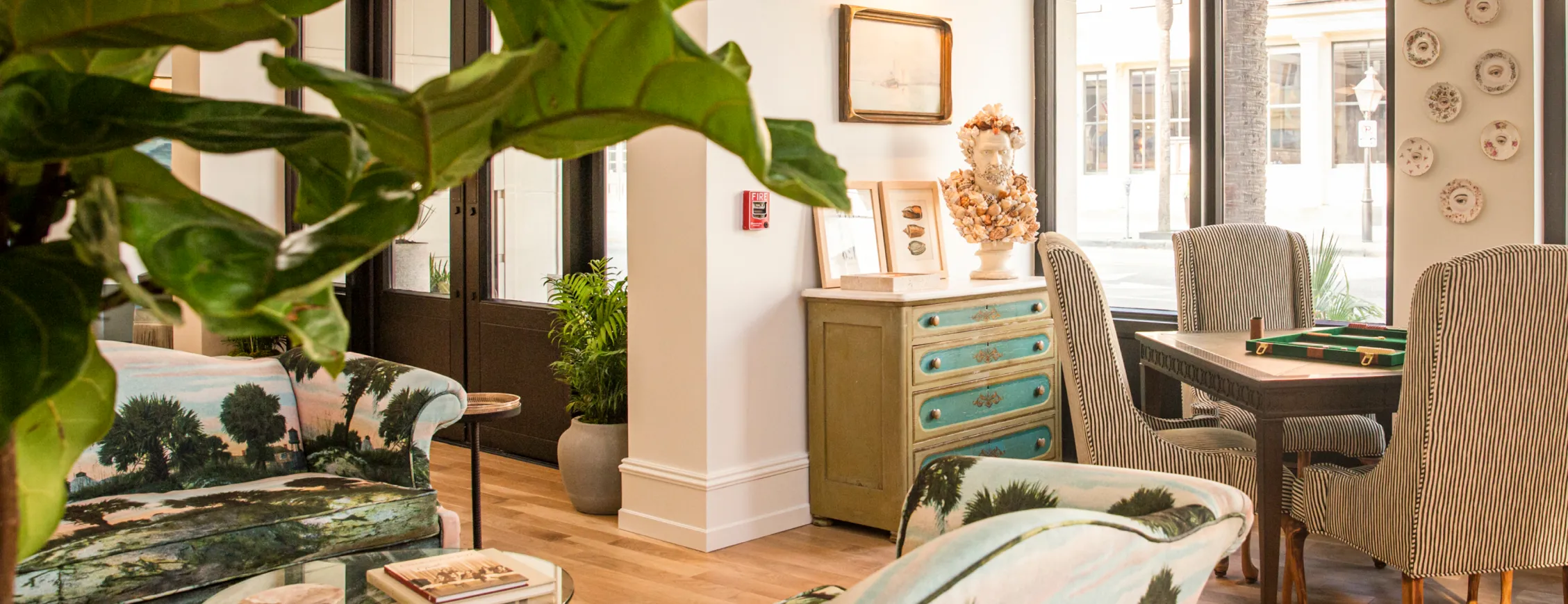 Bright living room with tropical print sofas, a green vintage dresser topped with a decorative bust, striped dining chairs around a wooden table, and large leafy plants.