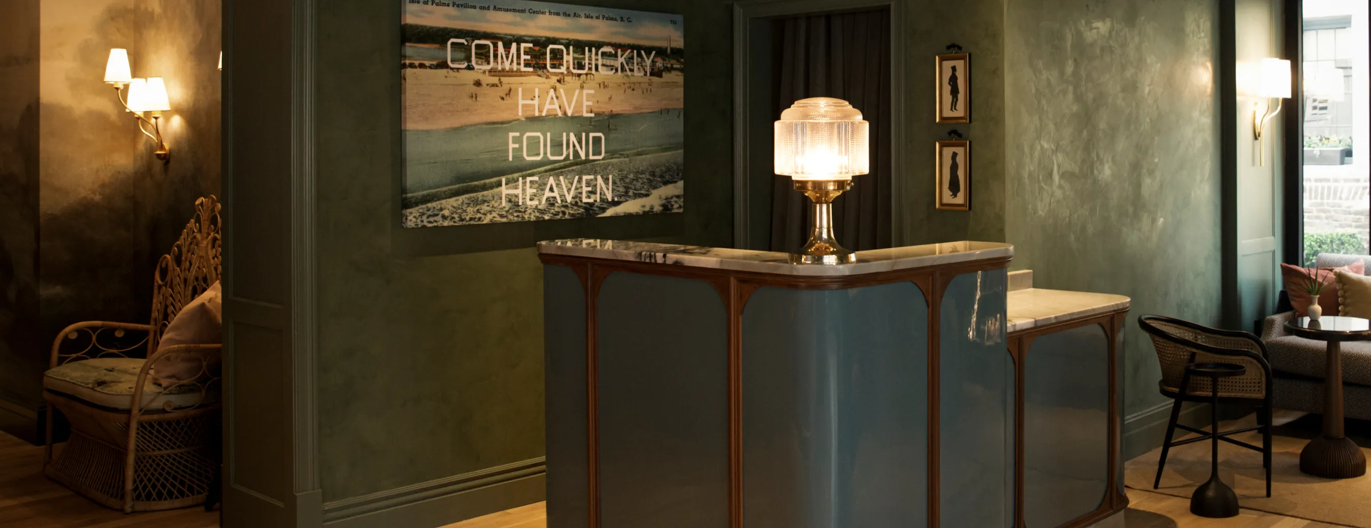 Cozy interior with a blue reception desk topped with a vintage lamp, wicker chairs, wall sconces, and a wall art that reads 'COME QUICKLY HAVE FOUND HEAVEN'.