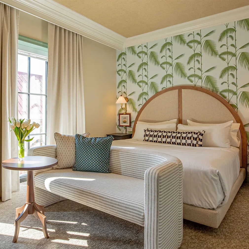 Bedroom with a round-arched upholstered headboard, tropical leaf wallpaper, a striped loveseat, side table with vase of white tulips, and glass doors with beige curtains leading to a balcony.