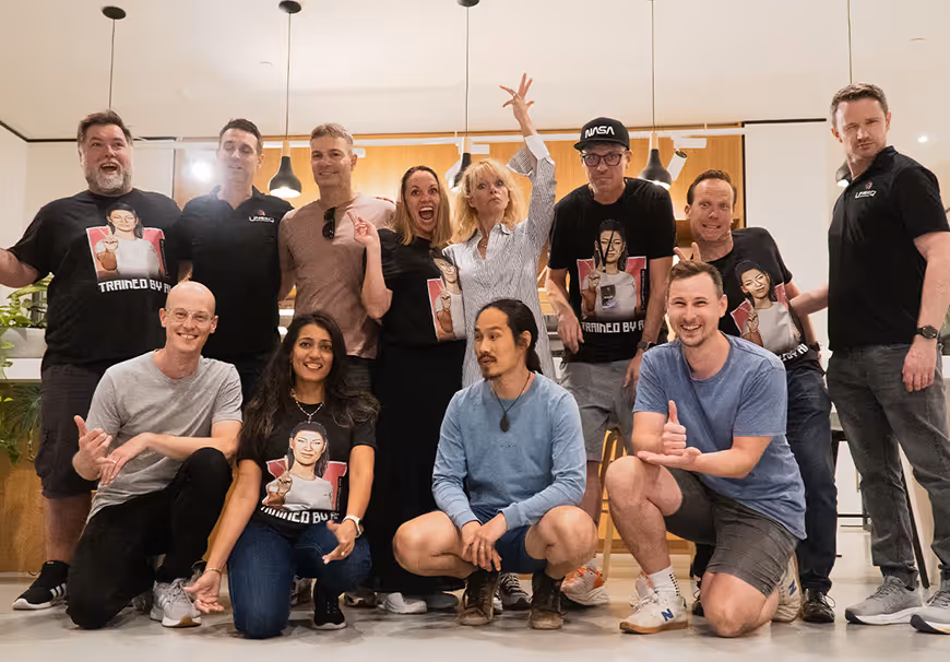 Group of twelve people posing indoors, some wearing matching black t-shirts with a graphic and text, smiling and making playful gestures.