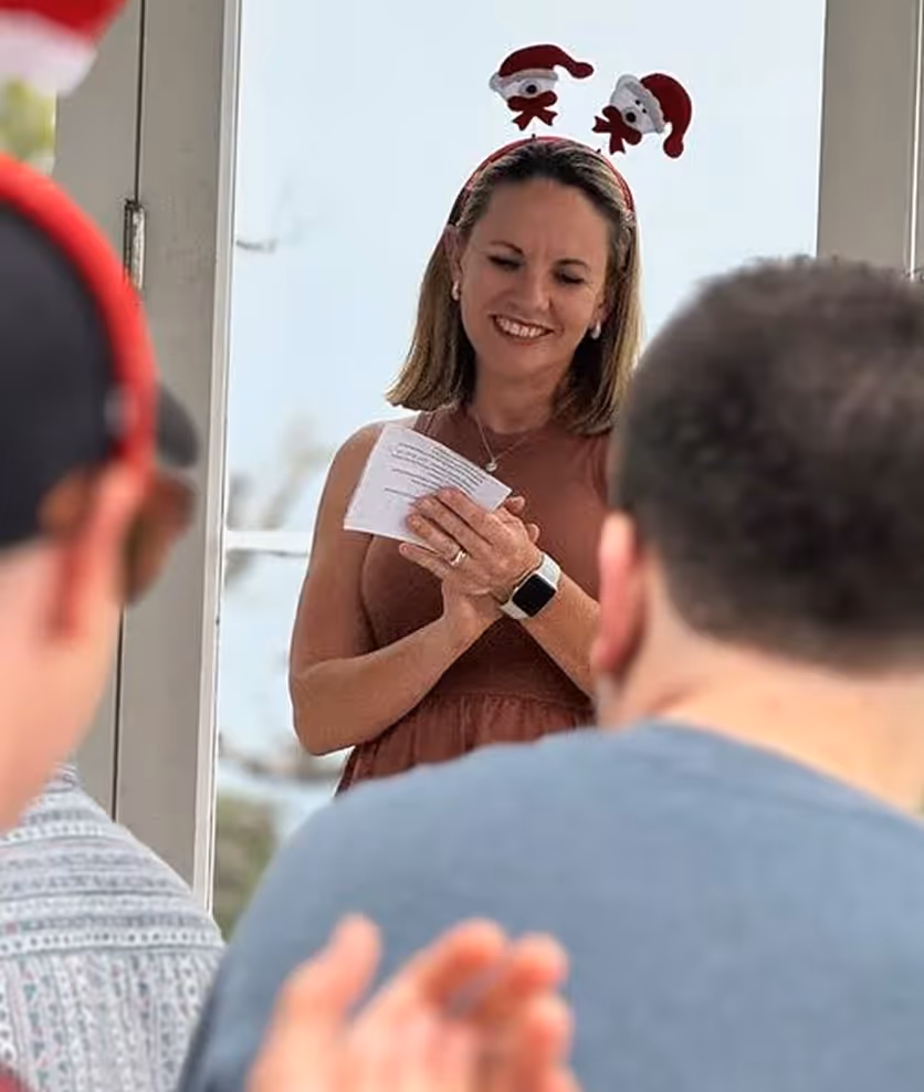 Smiling woman wearing a headband with Santa figures, holding papers and applauded by people.