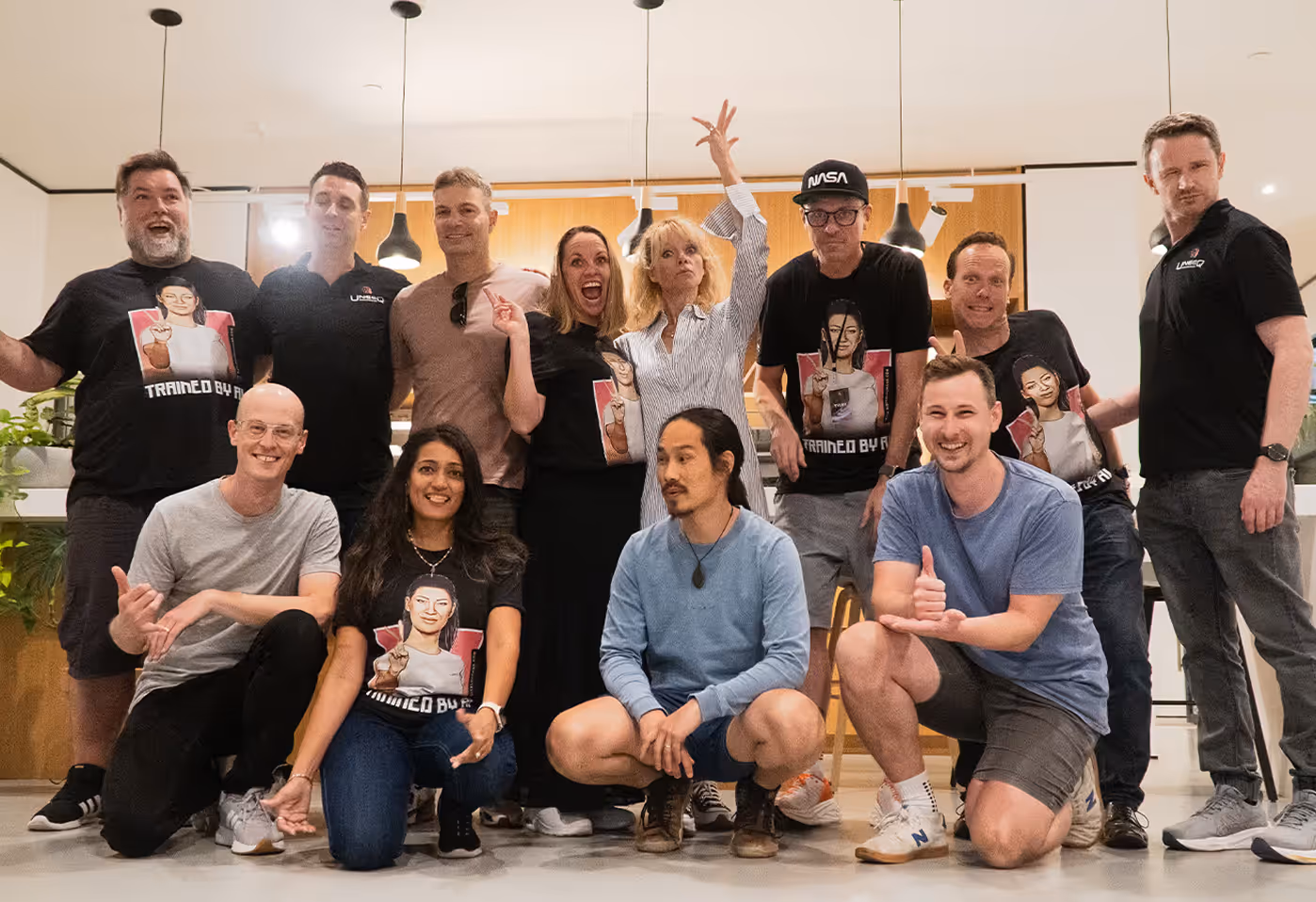 Group of twelve diverse adults posing cheerfully indoors, some wearing black T-shirts with a graphic design and the text 'TRAINED BY AI'.