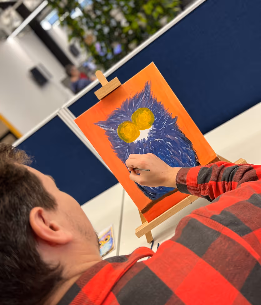 Person wearing a red and black checkered shirt painting a blue owl with large yellow eyes on an orange canvas.