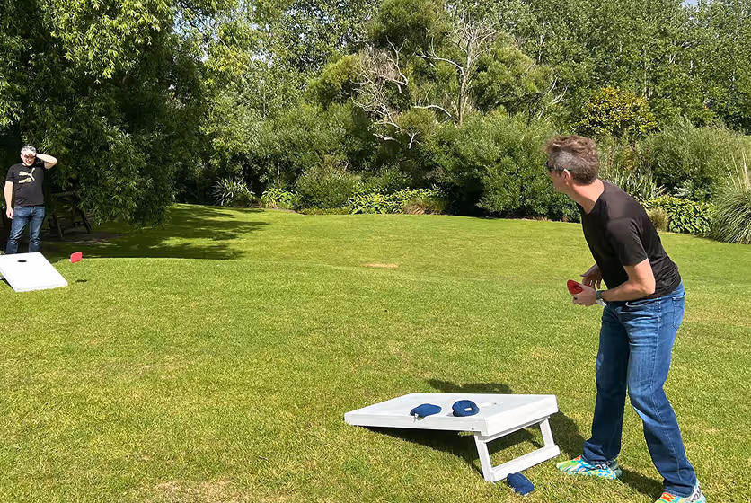Two men playing cornhole on a grassy lawn surrounded by trees, one preparing to toss a red bean bag while the other watches near the opposite board.