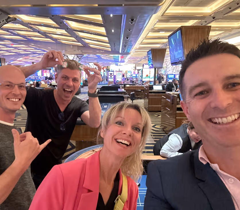 Four smiling adults taking a selfie together inside a lively casino with gaming tables and slot machines in the background.