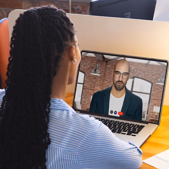A woman with braided hair wearing a blue and white striped shirt participates in a video call on a laptop with a bald man wearing glasses and a blazer, set against a brick wall background.