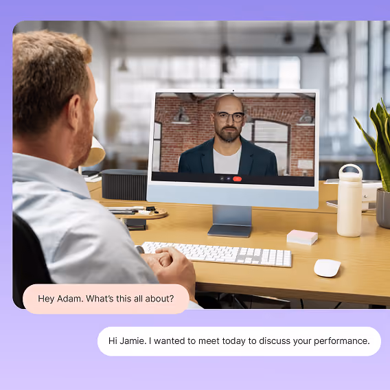 A man with short hair sits at a desk looking at a computer screen during a video call with a bald man wearing glasses and a dark blazer against a brick wall background; chat bubbles read: 'Hey Adam. What's this all about?' and 'Hi Jamie. I wanted to meet today to discuss your performance.'