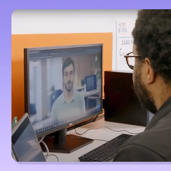 Man with glasses viewed from behind, sitting at a desk looking at a computer monitor displaying a video call with another man wearing a light green shirt.