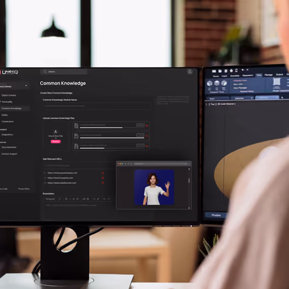 Two computer monitors on a desk showing a digital interface; the left screen displays a 'Common Knowledge' module with options to upload files, add URLs, and write descriptions, while the right screen shows a 3D modeling application. A person is partially visible in the foreground.