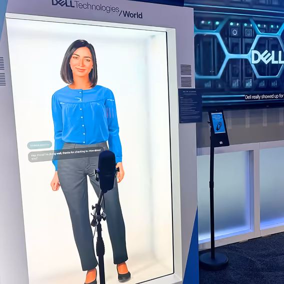 A life-like digital human in a blue blouse and gray pants displayed inside a white booth with 'Dell Technologies World' branding above it, next to a microphone and a screen showing the Dell logo.