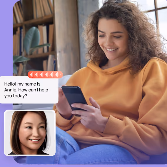 A smiling woman in a yellow hoodie is sitting and looking at her smartphone, engaging in a video call with a woman named Annie who appears in a small window and says, 'Hello! My name is Annie. How can I help you today?'.