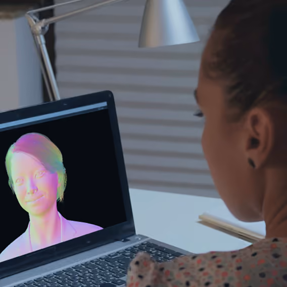 Person with pulled-back hair sitting at a desk, viewing a laptop screen that displays a colorful 3D-rendered head of a woman on a black background.