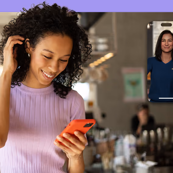 Smiling woman with curly hair wearing a purple sweater looking at her orange smartphone, with a blurred café background and a small inset of a video call featuring another woman.