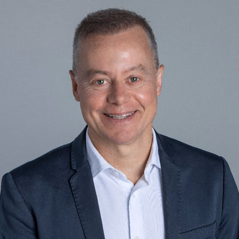 Portrait of a smiling man with short hair wearing a dark blazer over a white shirt against a gray background.