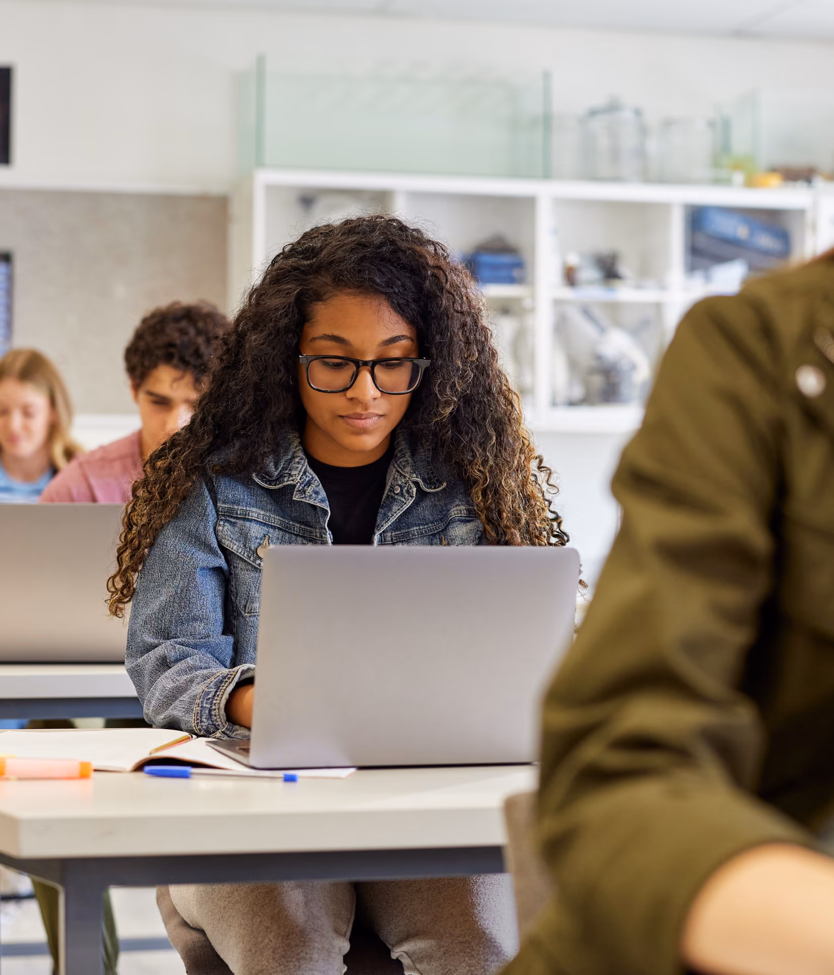 College student with long curly hair and glasses sits at a desk focused on her open laptop, with other students working on laptops in the background in a classroom setting.