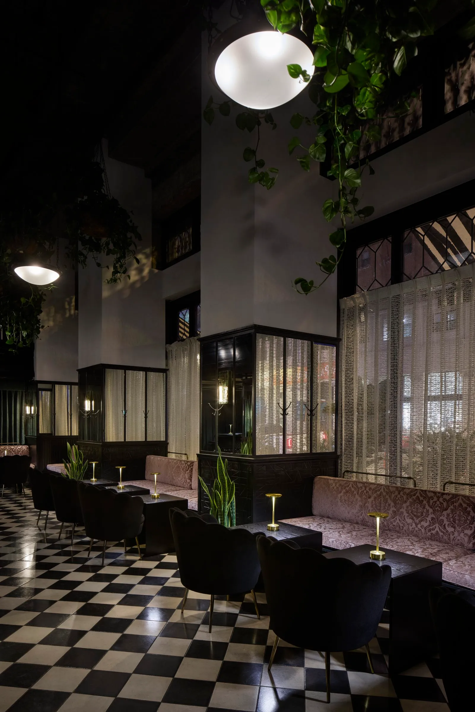 Pink damask banquettes and black scalloped chairs beside windows with lace curtains at Mary at the Stile Hotel at night.