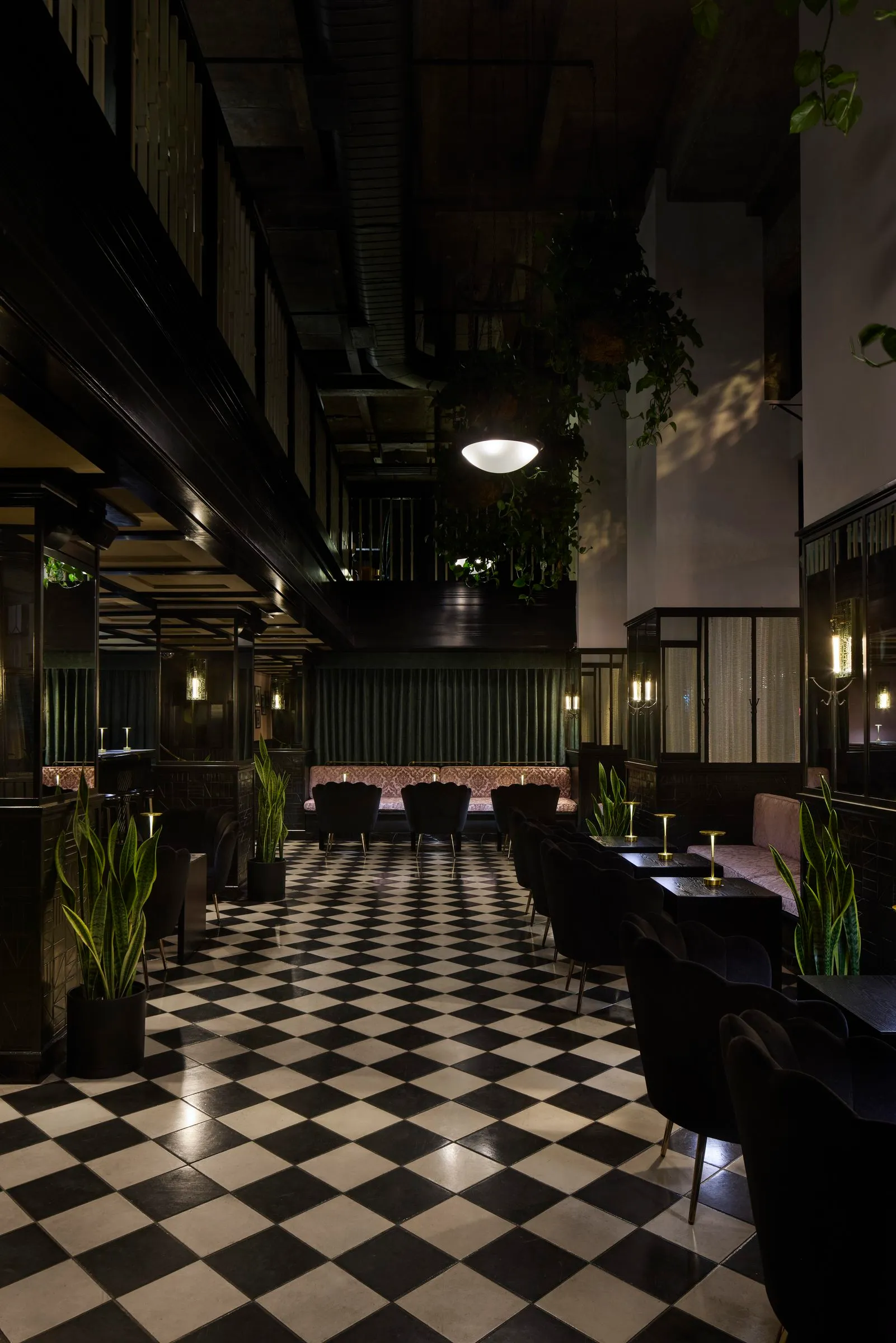 Hanging plants and a pendant light above black scalloped chairs and checkered tile at Mary at the Stile Hotel at night.