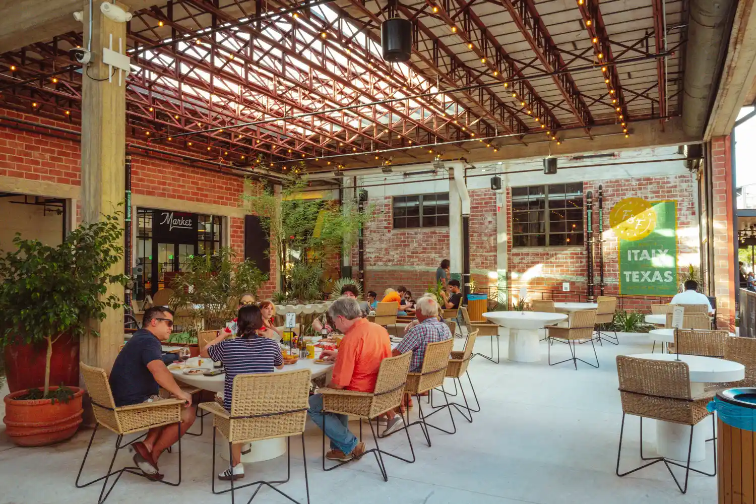 Outdoor restaurant seating with patrons at tables inside a courtyard with brick walls and overhead string lights