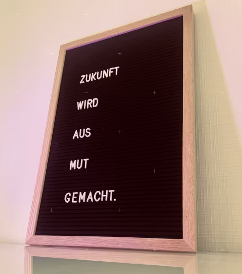 Ein heller Holzrahmen mit schwarzem Innenteil zeigt in weiß gesetzten Buchstaben den Spruch: Zukunft wird aus Mut gemacht