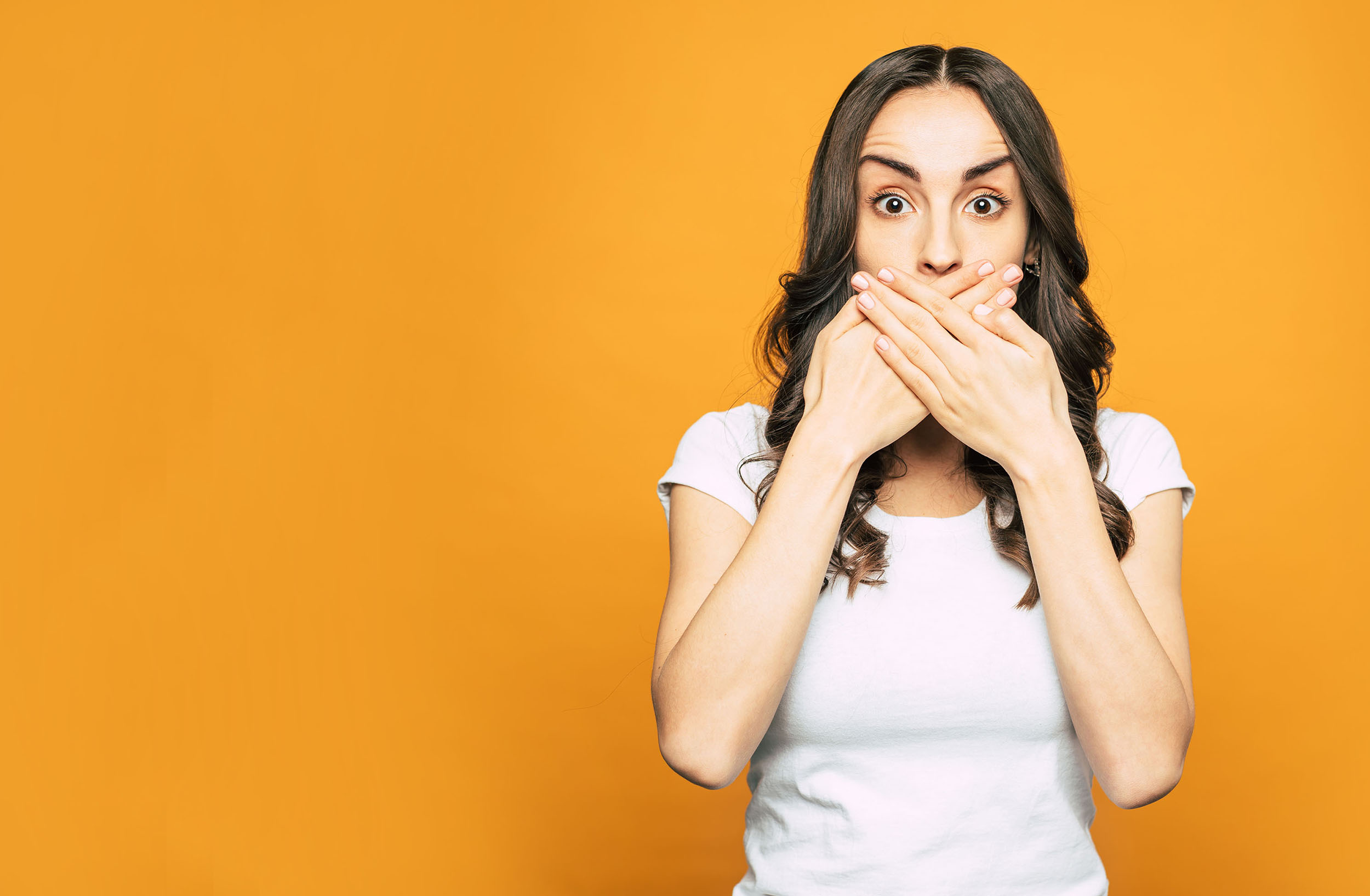 A nervous-looking woman with her hands over her mouth against a plain background