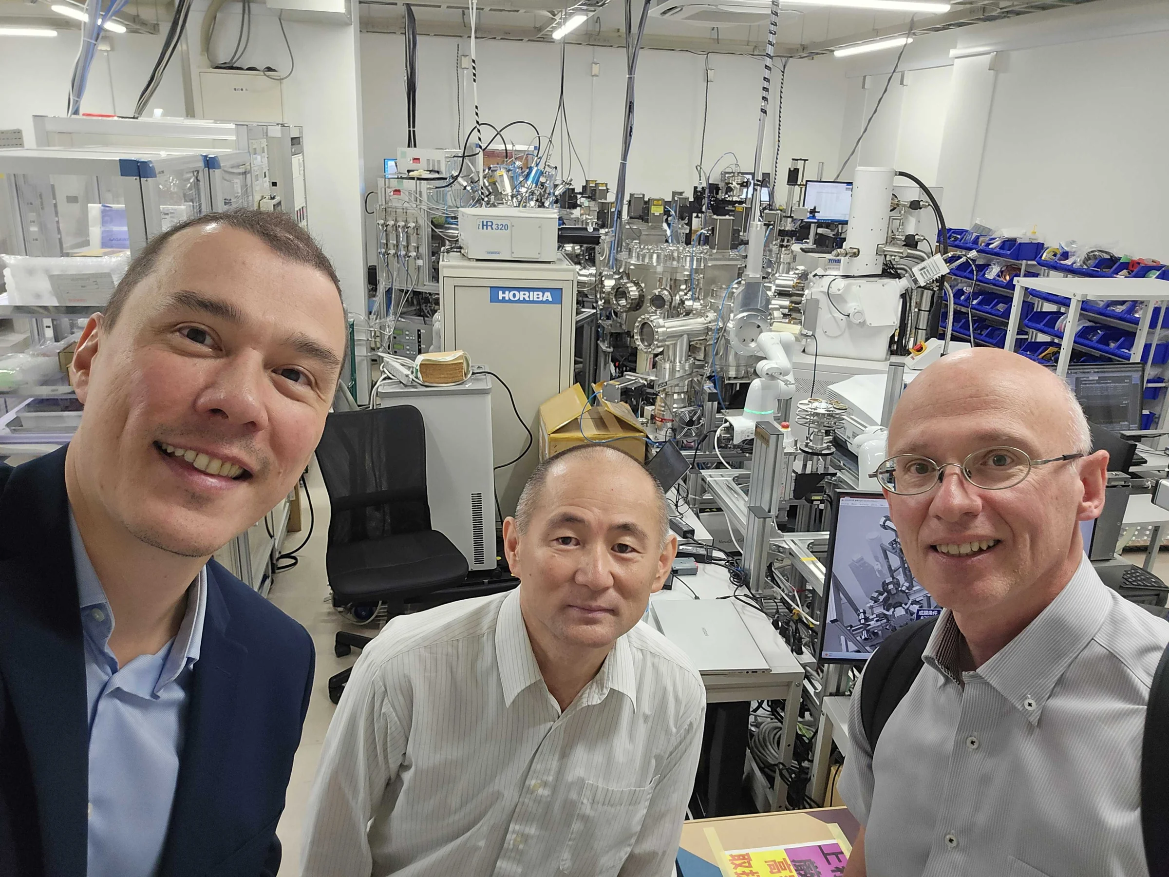 研究室の実験装置の前で撮影した、Mat3raチームのメンバー2名と担当者の自撮り写真