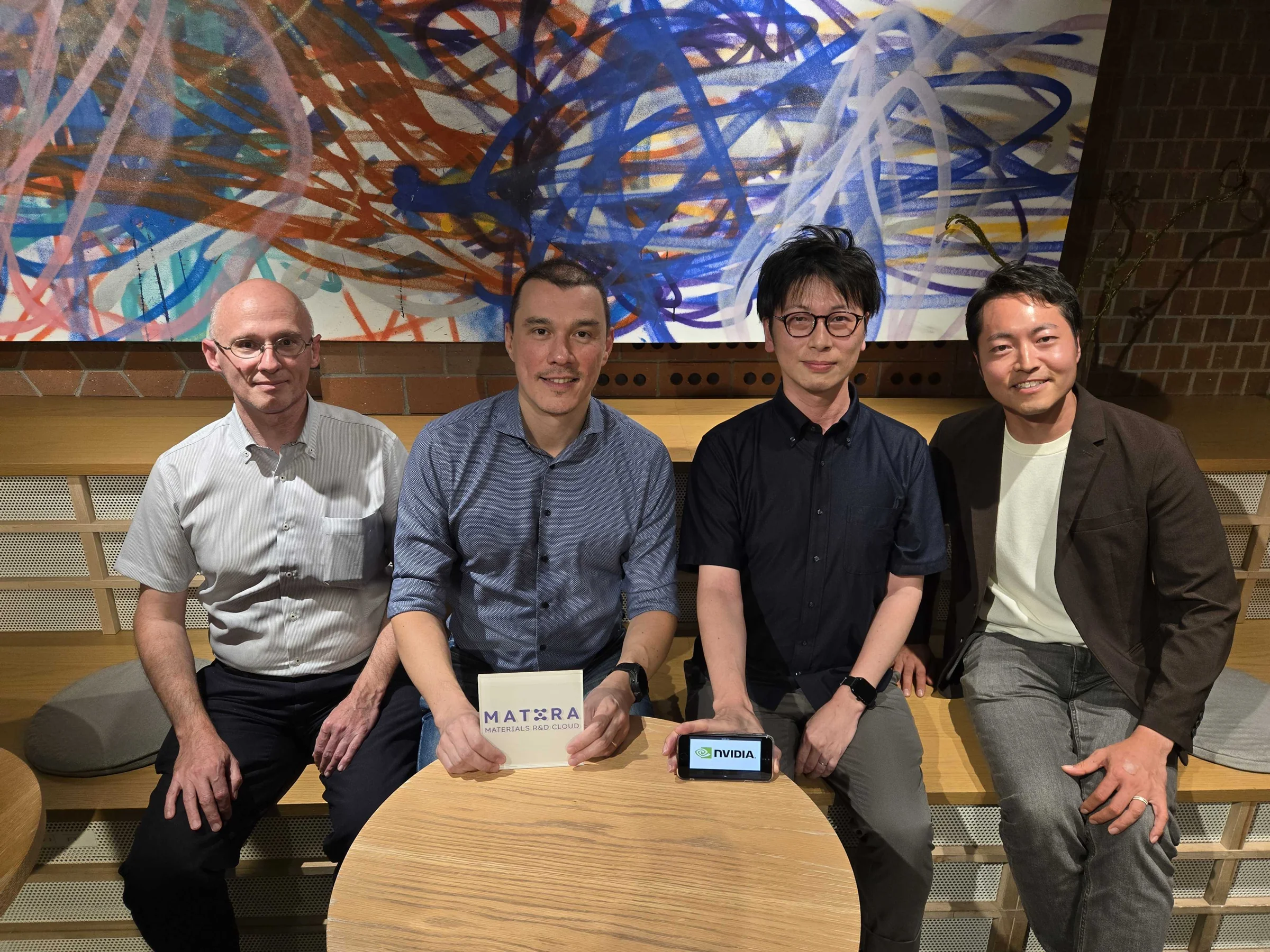 Two members of the Mat3ra team and two hosts seated at a wooden table, holding a Mat3ra Materials R and D Cloud block and a device showing the NVIDIA logo