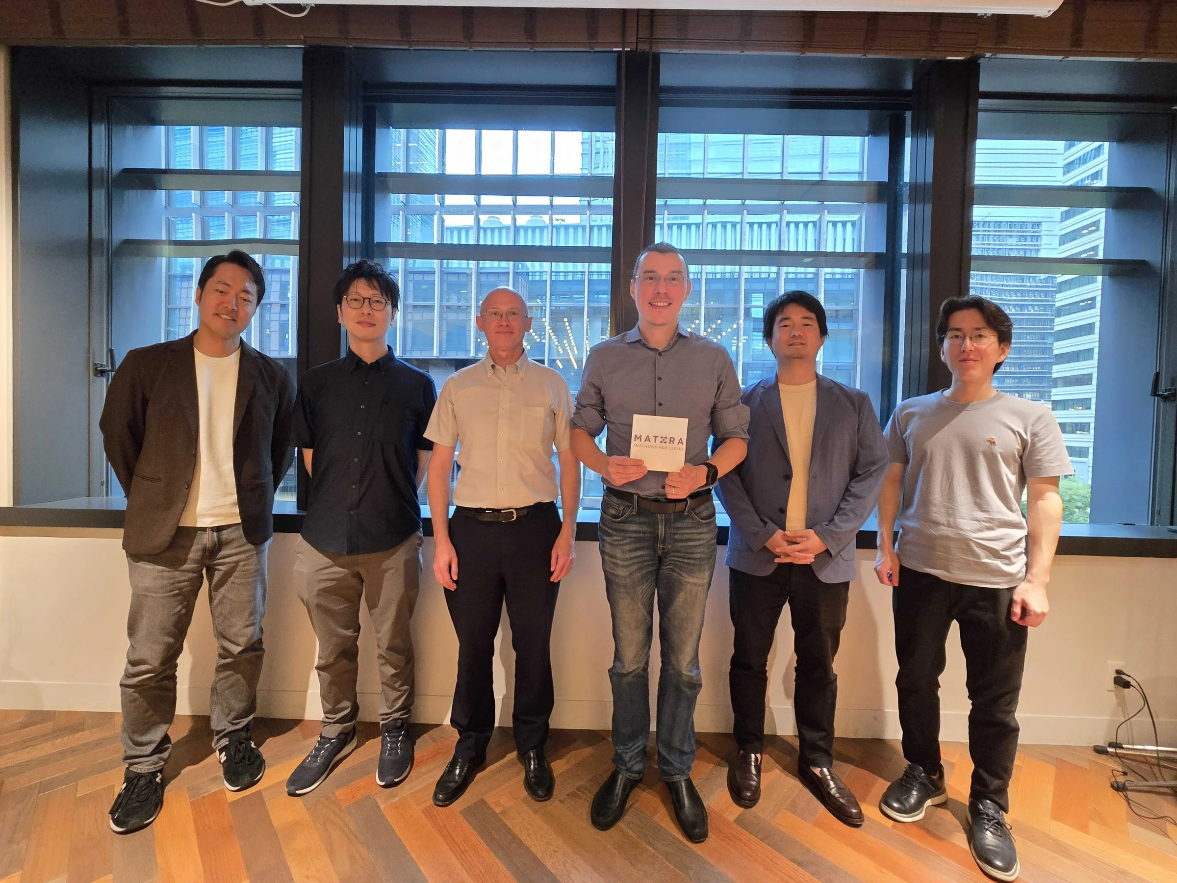 Group photo of the Mat3ra team and hosts in a meeting room with a city view, holding the Mat3ra Materials R and D Cloud block
