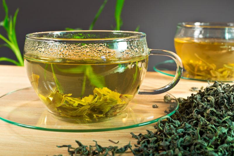 Glass cup of freshly brewed green tea with loose tea leaves, sitting on a clear saucer next to a pile of dried green tea leaves.