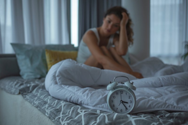 A person sits on a bed with an alarm clock in the foreground, appearing distressed.