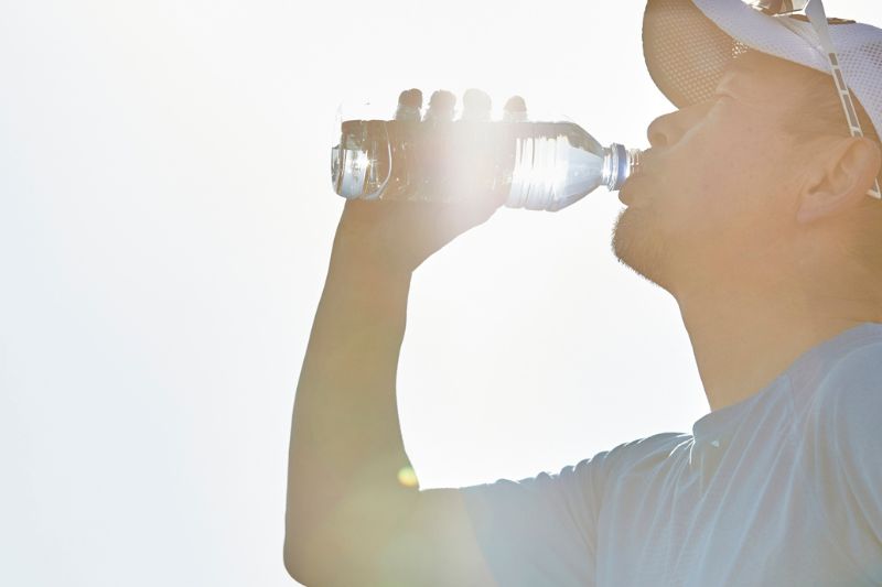 A person wearing a white cap and blue shirt drinks from a clear plastic water bottle against a bright, sunlit sky.