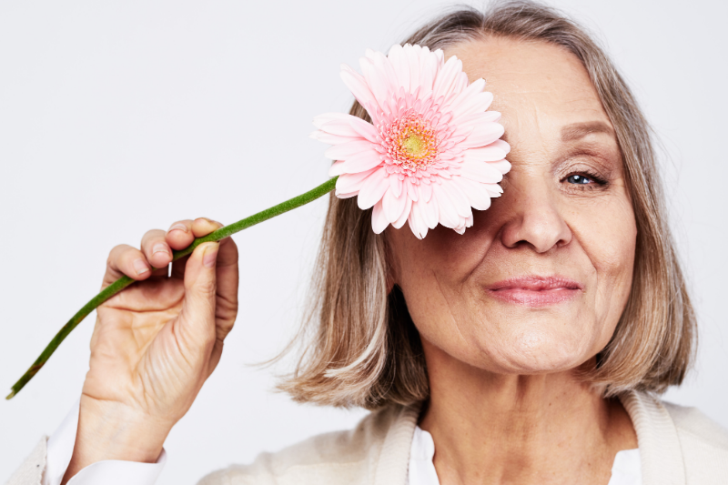 A person holds a pink gerbera daisy in front of their right eye against a plain background.