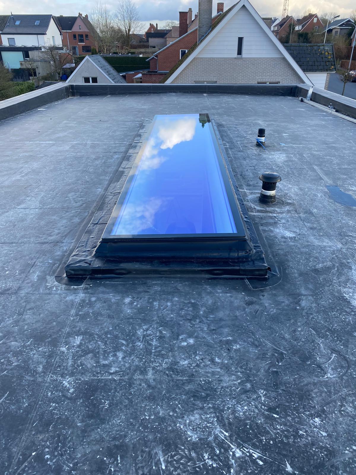 Rectangular skylight window on a flat black rooftop reflecting blue sky and clouds, with houses in the background.