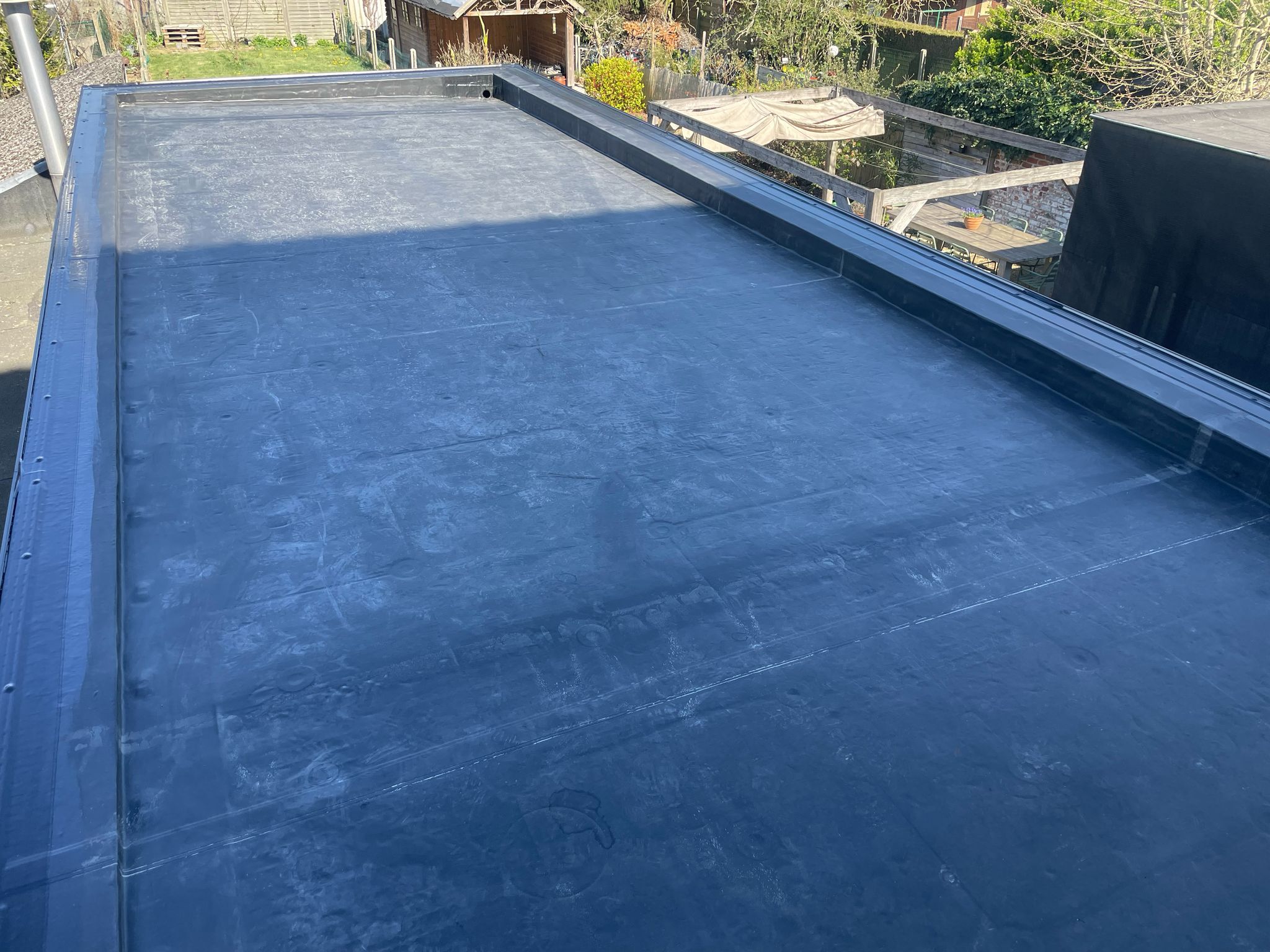 Flat rubber membrane roof on a building with garden and outdoor seating area in the background.
