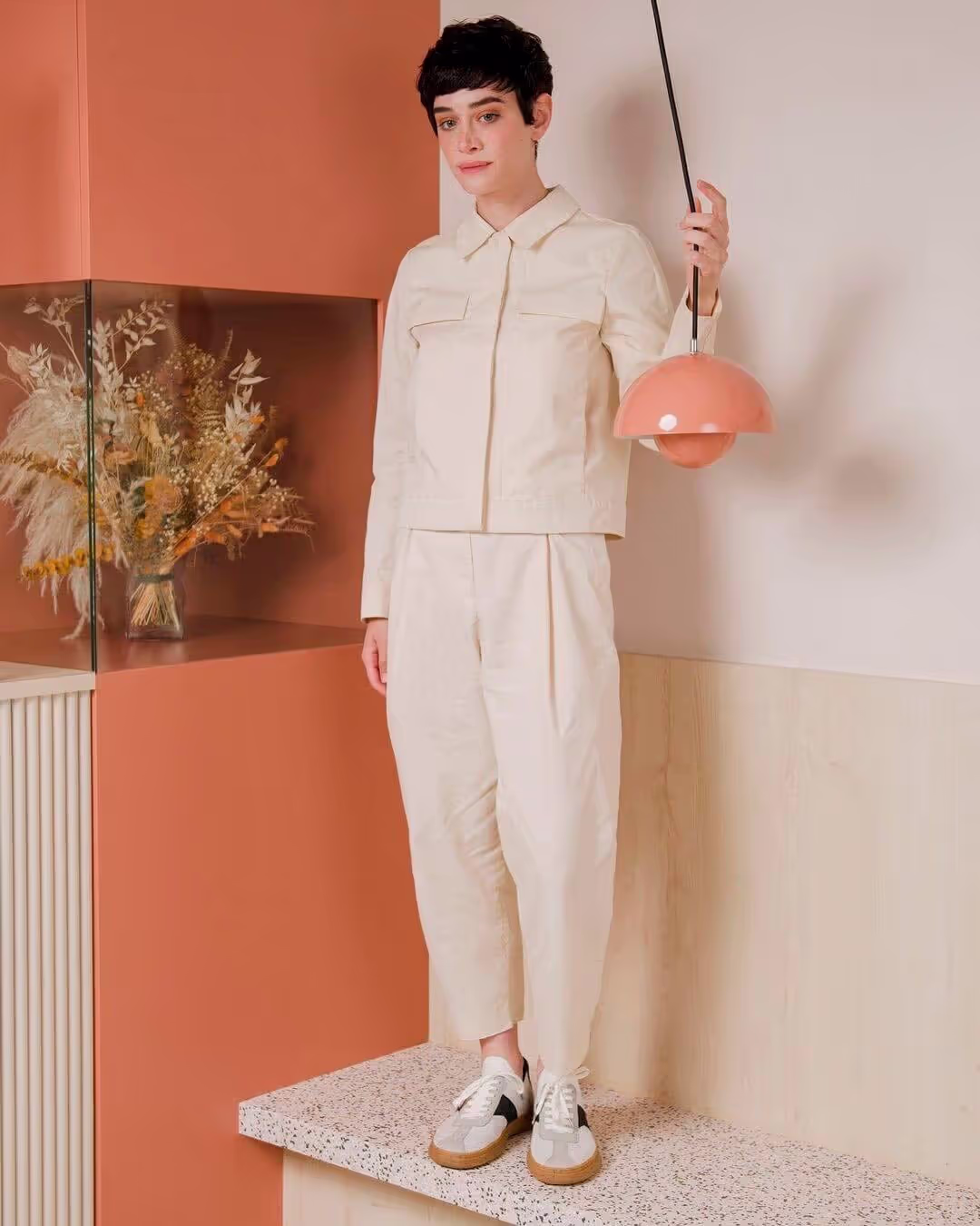 A model in a minimalist beige outfit stands on a terrazzo platform, holding a suspended peach-colored pendant light in a modern, earth-toned interior