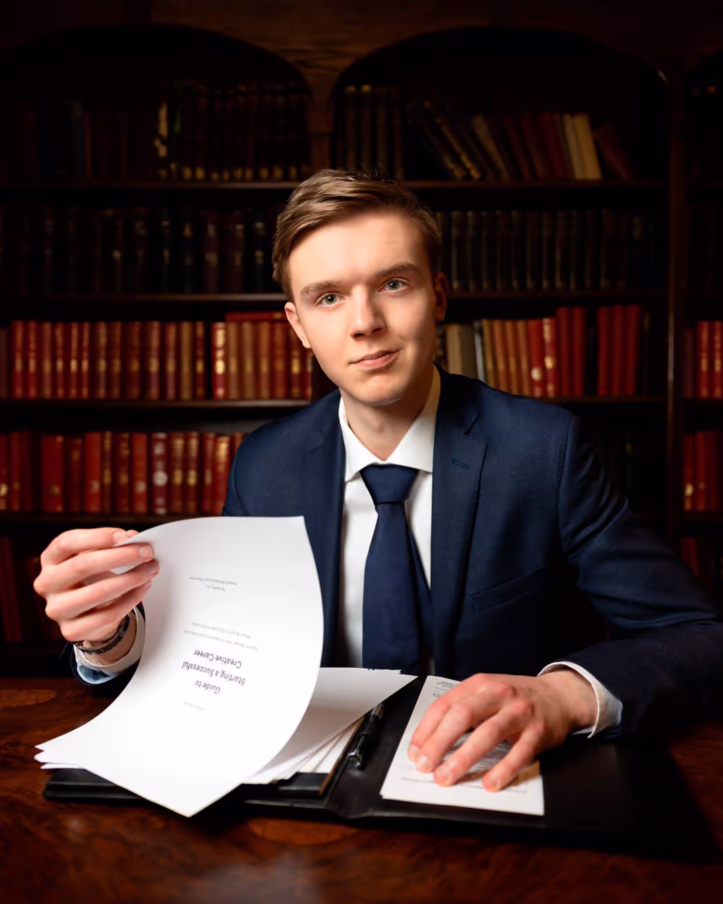 Andrew Reutsky in a navy blue suit, sitting at a wooden desk in a library, holding a printed guide titled “Guide to Starting a Successful Creative Career.”