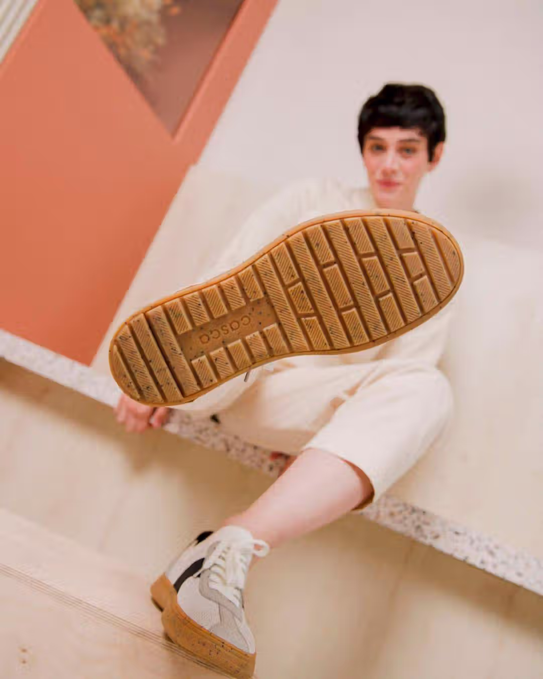 A model in a minimalist beige outfit sits on a terrazzo platform, holding a suspended peach-colored pendant light in a modern, earth-toned interior