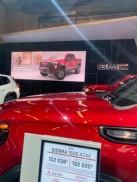 A red GMC Sierra 1500 AT4X is shown at an auto show with a large display screen, pricing sign, and other GMC vehicles around it.