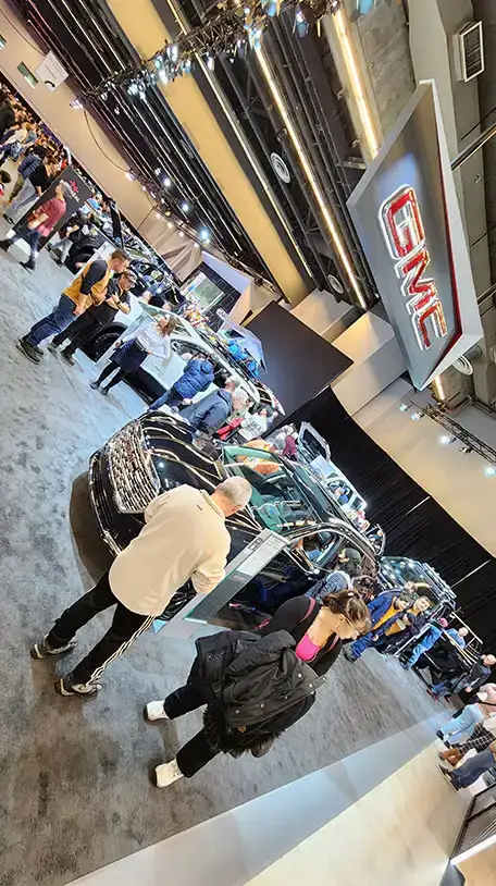 Visitors walk through a GMC auto show with multiple vehicles on display, a large GMC sign overhead, and a black car in the foreground.