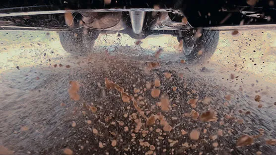 The rear wheels of a vehicle are driving through a puddle, sending up dramatic splashes of muddy water in multiple directions. The vehicle body is partially visible at the top of the frame.